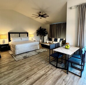 Habitación de hotel con cama, ventilador de techo, mesa de comedor con sillas, planta en maceta y mesita de noche con lámpara.