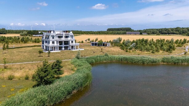 Una casa moderna de dos pisos con un balcón grande, rodeada de campos y un estanque.