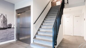 Una escalera con barandilla de metal, una puerta con número y una pared con un mural de paisaje urbano.