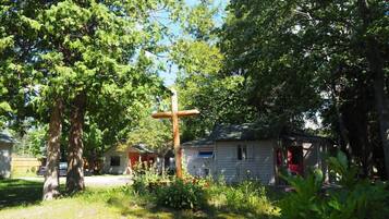 A rustic cabin with a wooden cross in a lush garden, surrounded by trees and a small shed.