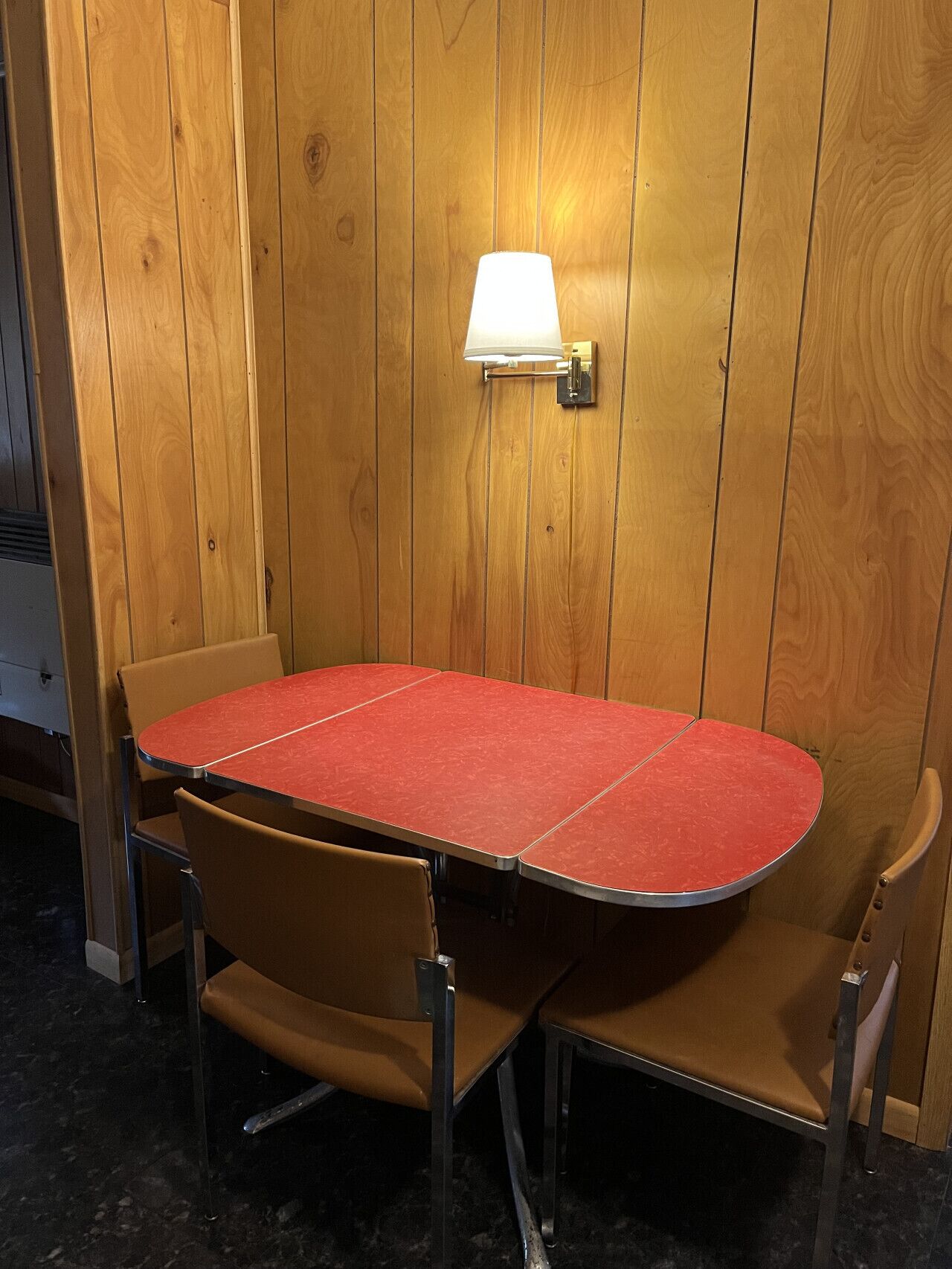 A wooden-paneled room with a red table and four brown chairs. A wall-mounted lamp is visible.