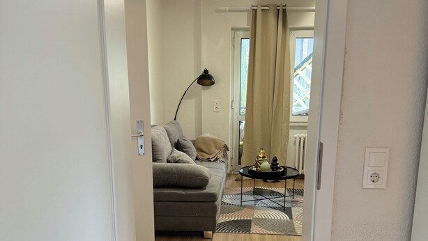 A living room with a sofa, a lamp, a patterned rug, and a view of the outside through a window with curtains. - Be Home in Dortmund (Dortmund)