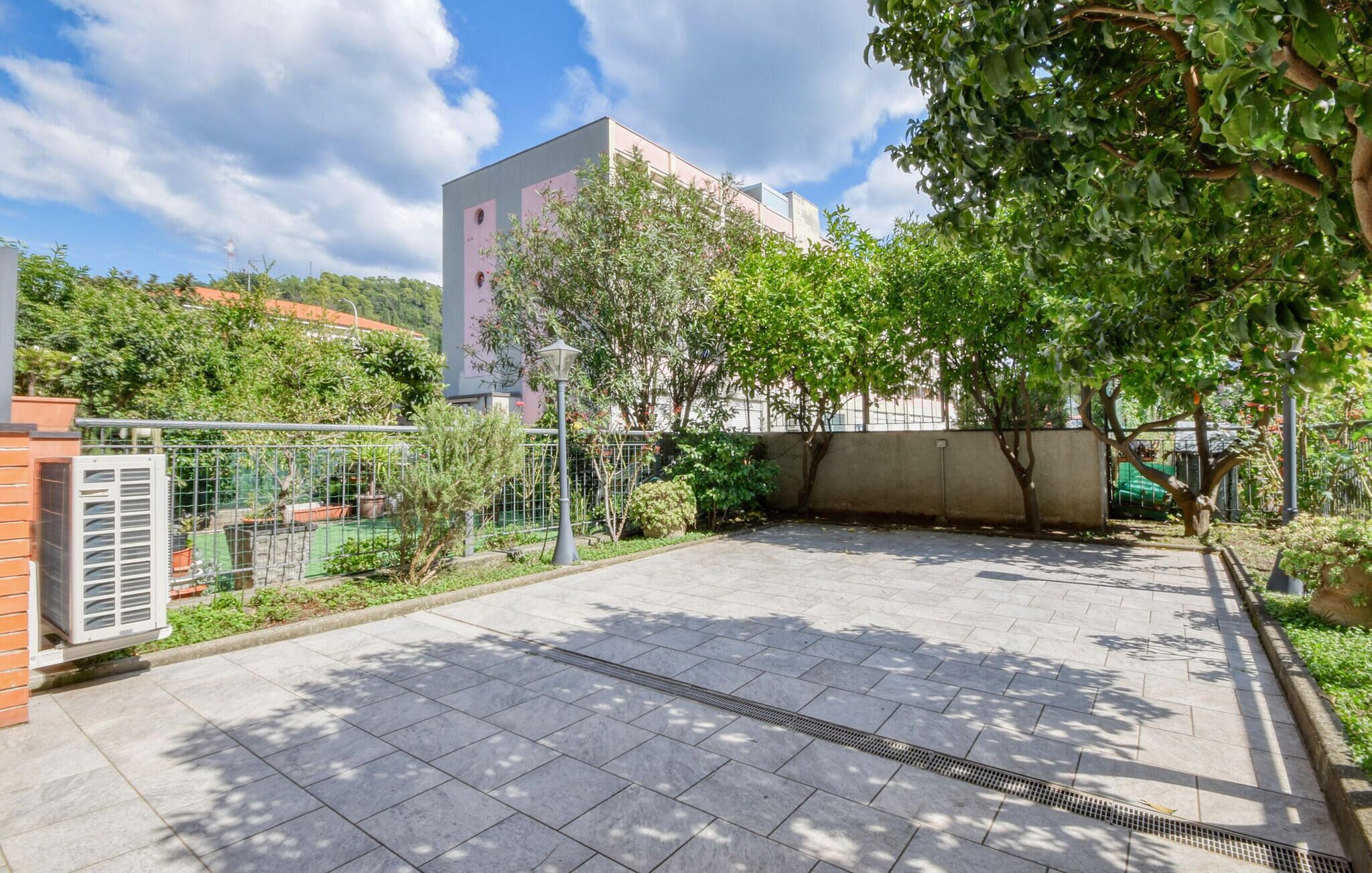 An outdoor courtyard with a paved floor, surrounded by greenery and a metal fence.