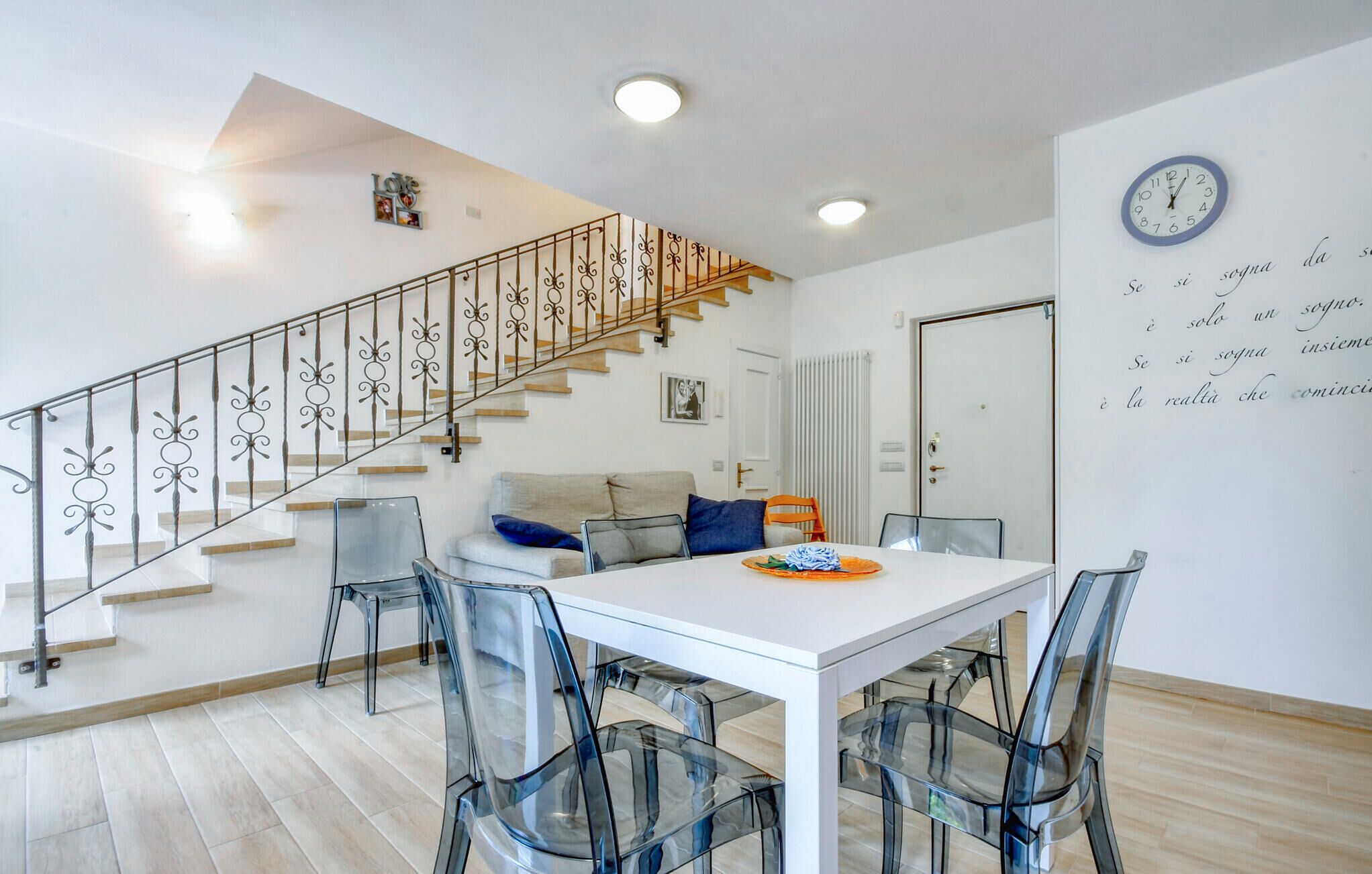 Un espace de vie moderne comprenant un escalier, un coin repas avec des chaises transparentes, un canapé et une horloge murale.