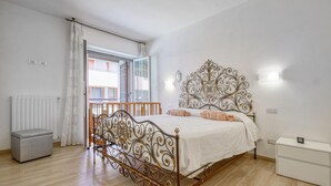 A bedroom with a bed, a radiator, a window with curtains, a bedside table, and a small cabinet.