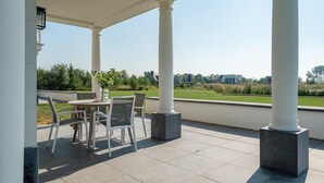 A covered patio area with a table and chairs, large columns, and a view of a green landscape and buildings in the distance.