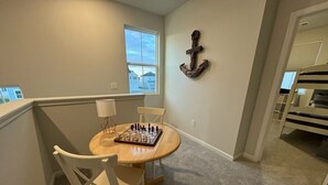 Une pièce avec une table ronde en bois, deux chaises blanches, un jeu d’échecs, une lampe et, au fond, un lit superposé.