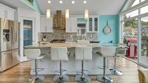 A modern kitchen with a central island, bar stools, stainless steel appliances, and a large window offering a view of the outside. - La Casa Cottage Beach Vibe, Entire Home Lake Views (Kelowna)