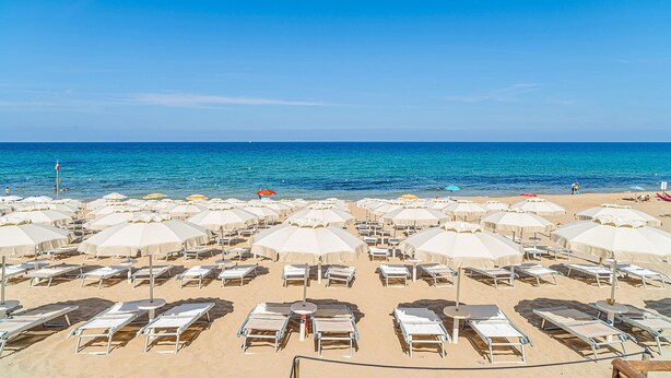 Ein Strand mit Liegestühlen und Sonnenschirmen, einem klaren blauen Himmel und dem Meer im Hintergrund.