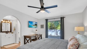 A bedroom with a bed, a desk, a chair, a ceiling fan, a window with curtains, and a view of greenery outside. - City Center Townhouse with Private Yard! (Tucson)