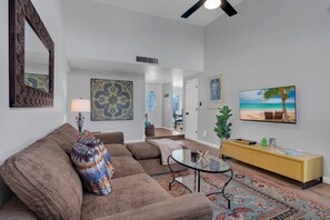 A living room with a brown sofa, a glass coffee table, a yellow console table, and a large beach-themed TV. - City Center Townhouse with Private Yard! (Tucson)