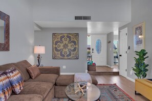 A living room with a brown sofa, a glass coffee table, a patterned wall hanging, and a door leading to another room.