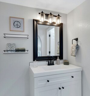 Une salle de bain avec un meuble-lavabo blanc, un grand miroir avec un cadre noir et un luminaire placé au-dessus du miroir. Deux porte-serviettes sont fixés au mur, l’un avec une serviette et l’autre avec une serviette et une bouteille.