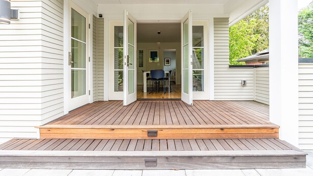 En overdækket veranda med træterrasse, hvid facade og udsigt ind til huset.