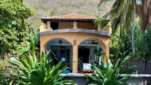 A two-story building with a tiled roof, a covered patio area, and a garden with large green plants. - Casa Pelicanos is a private beachfront home with pool. Short walk to town.
 (Troncones)