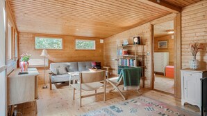 Een knus interieur van een chalet met een houten plafond, een bank, een eettafel, een boekenkast en een kleine keukenhoek.