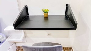 Ein Zimmer mit einem an der Wand befestigten, schwarzen Metallregal, einer kleinen Topfpflanze auf dem Regal, einem grauen Polsterstuhl und einem weißen Nachttisch.
