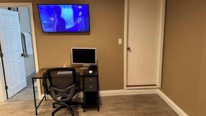 Ein Zimmer mit Schreibtisch aus Holz, einem Computer, einem Stuhl und einem an der Wand befestigten Fernseher.