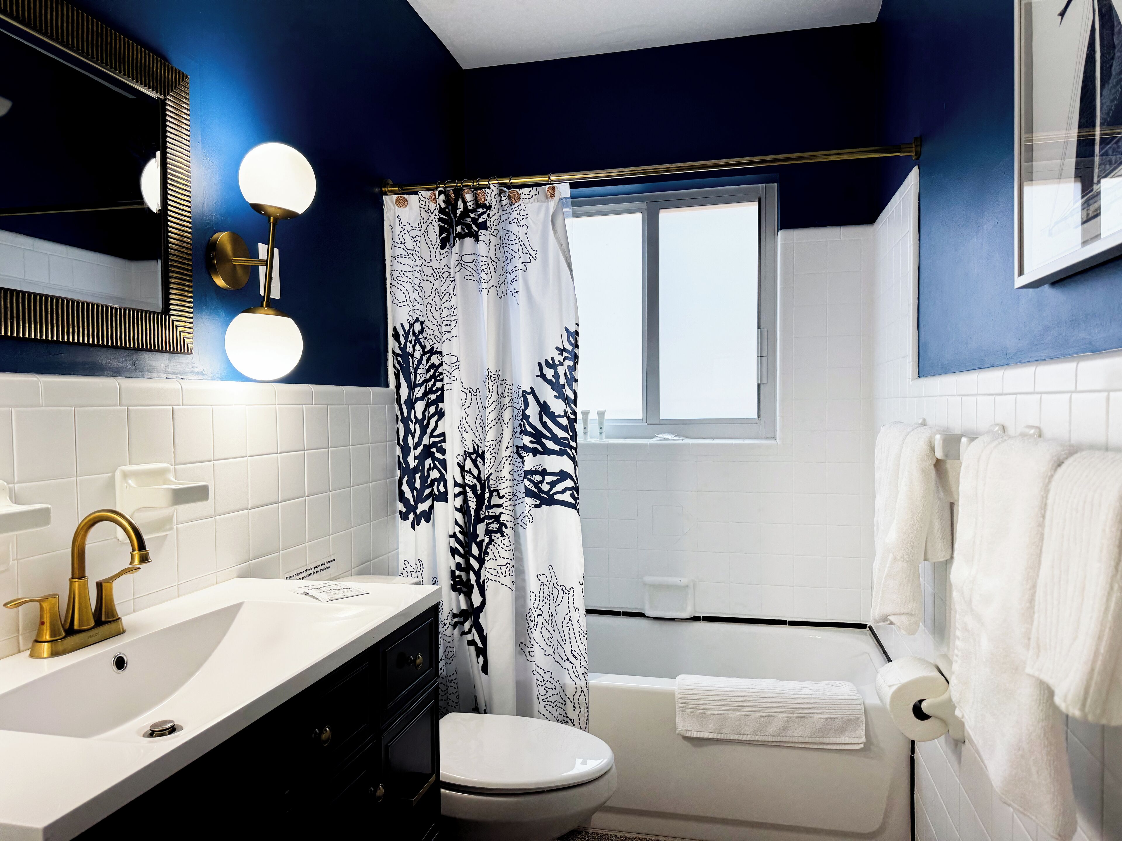 Une salle de bain avec un lavabo blanc, un robinet doré et des toilettes blanches. Il y a une fenêtre avec un rideau blanc, et des serviettes blanches sont accrochées au mur.