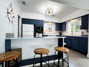 Uma cozinha moderna com armários azul-escuro, geladeira branca, uma ilha de vidro e dois banquinhos de madeira.