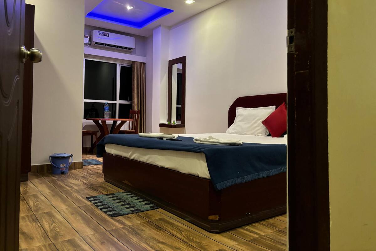 A hotel room with a bed, a desk, a chair, a window, and an air conditioning unit.