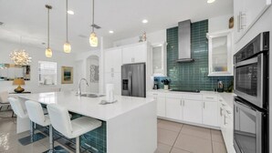 Een moderne keuken met witte kasten, een centraal kookeiland met barstoelen, roestvrijstalen apparatuur en hanglampen.