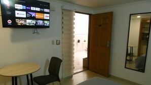A hotel room with a TV displaying various streaming services, a wooden door, a small table, a chair, and a mirror. - BogoRooms Héroes  (Bogotá)