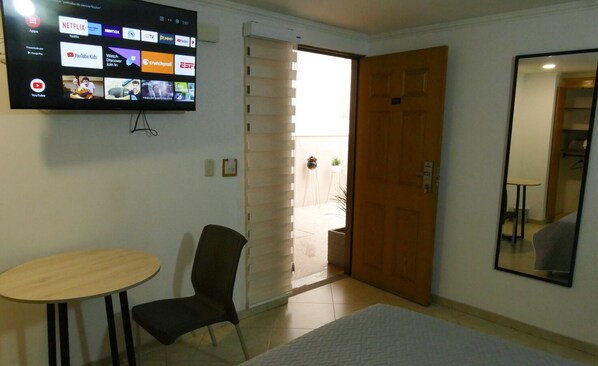 Habitación de hotel con televisión que muestra varios servicios de streaming, una puerta de madera, una mesita, una silla y un espejo.