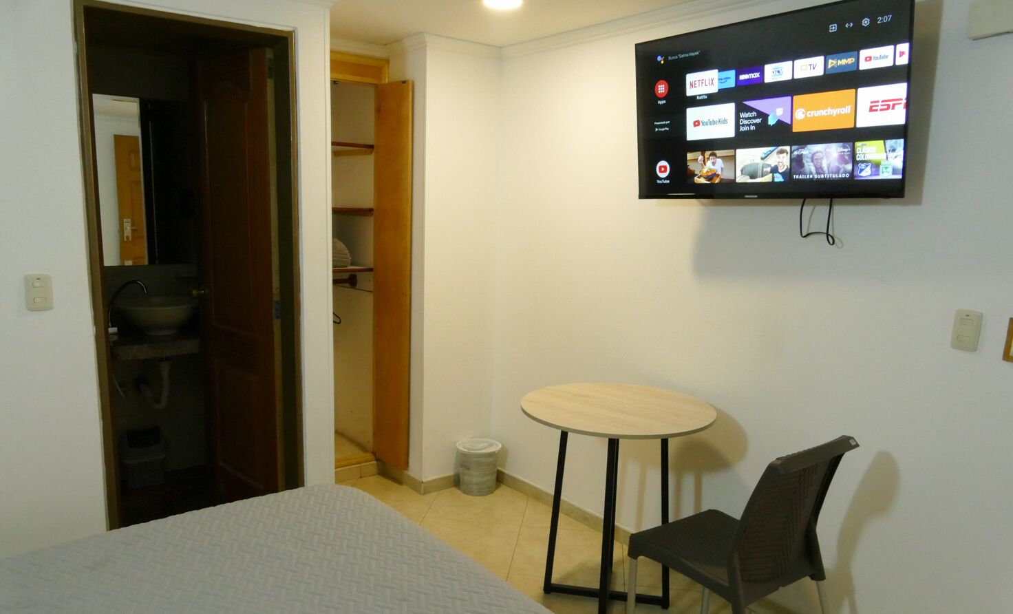 Una habitación con cama, una mesita, una silla, un televisor fijado en la pared que muestra varios servicios de transmisión, y un armario con estantes.