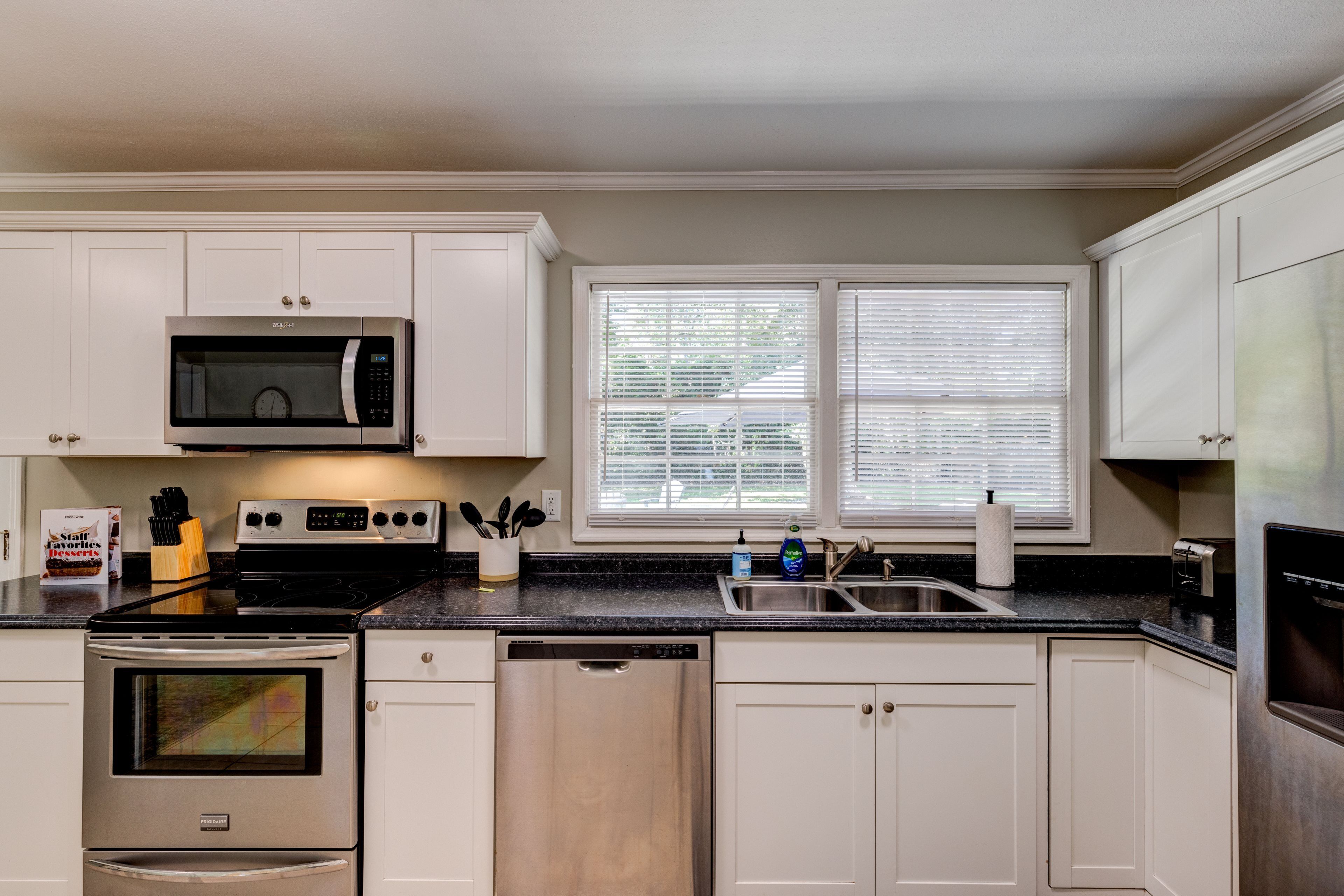 A modern kitchen with white cabinets, stainless steel appliances, a dishwasher, and a window with blinds.