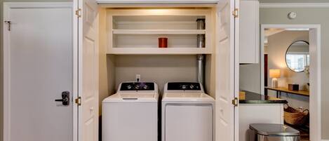 A laundry room with two front-loading washers and dryers, a trash can, and a shelf with a red cup.