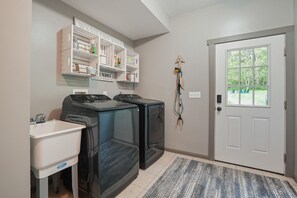 A laundry room with two front-loading washers, a white sink, and shelves above containing cleaning supplies. - Walnut Hill Lodge | Hocking Hills Luxury, Hot Tub (Logan)