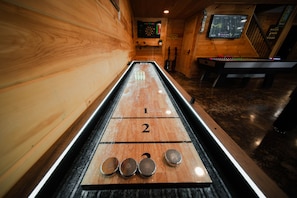 A shuffleboard table with numbered scoring areas, a dartboard, and a pool table in a wooden room.