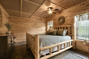 A wooden cabin bedroom with a bed, a dresser, a ceiling fan, and a window with curtains.