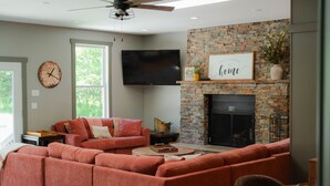 A living room with a sectional sofa, a fireplace, a flat-screen TV, and a clock on the wall. - Walnut Hill Lodge  | Hocking Hills Luxury, Hot Tub (Logan)
