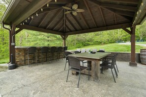 Un patio al aire libre con una zona de barbacoa de piedra, una mesa con sillas y vistas a un paisaje cubierto de césped.