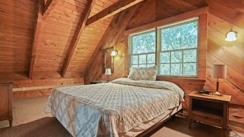 A cozy attic bedroom with a wooden sloped ceiling, a large window with blinds, a bed with a quilted bedspread, and a nightstand with a lamp.