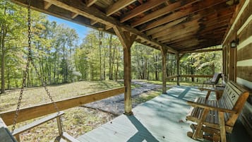A covered wooden deck with a swing, benches, and a view of a forest.