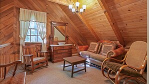 A cozy attic room with wooden walls, a wooden ceiling, a chandelier, a sofa, a rocking chair, a wooden table, and a dresser with a mirror.