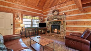 A cozy log cabin interior with a stone fireplace, leather sofas, and a television.