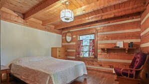 A wooden cabin room with a bed, dresser, chair, and a window with curtains.