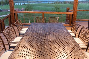 A covered outdoor seating area with a metal table and chairs, overlooking a grassy field and a lake.