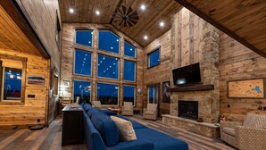 A log cabin interior with a long blue sofa, stone fireplace, and large windows. - All-Season Luxury MTN Retreat @ Lake w/ Hot Tub, Sauna & Near Hiking Trails, YNP (Island Park)