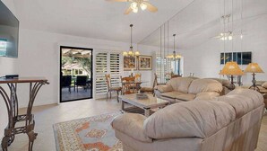 Uma sala de estar ampla com um sofá grande, uma mesa de centro e um ventilador de teto. É possível ver uma área de jantar através de uma porta de vidro.