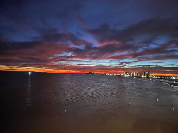 Auringonlasku meren rannalla, kaupungin siluetti näkyy horisontissa.
