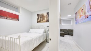 A bedroom with a white bed, a painting of the Eiffel Tower, a painting of a tree-lined path, and a painting of a woman in a dress. - Basement Suite in Vancouver (Vancouver)
