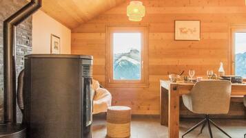 Ein gemütliches Blockhaus-Interieur mit Kamin aus Stein, Holzwänden, einer Essecke mit Tisch und Stühlen sowie einem Fenster mit Blick auf die Berge.