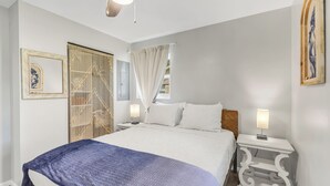 A bedroom with a bed, two bedside tables, a ceiling fan, a window with curtains, and a wall-mounted artwork. - Mermaid Cottage: 5 mi. from beach, fully stocked 3br, grill, deck, bunk bed (Fort Walton Beach)