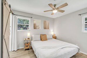 A bedroom with a bed, a ceiling fan, a window with curtains, a bedside table with a lamp, and wall art.