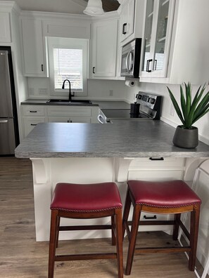 A kitchen with white cabinets, a granite countertop, a stainless steel refrigerator, a microwave, and a sink. There are two wooden stools with red cushions and a potted plant on the countertop.